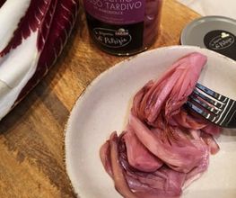 Eingelegter Radicchio in einer Schale mit Gabel, daneben ein Glas und frische Radicchio-Blätter.