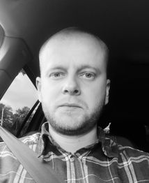 Mann mit Bart im Auto, schwarz-weiß Foto, trägt ein kariertes Hemd und Sicherheitsgurt.
