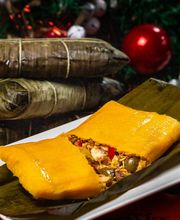 Hallaca venezolana sobre plato, rodeada de hojas verdes, con adornos navideños al fondo.