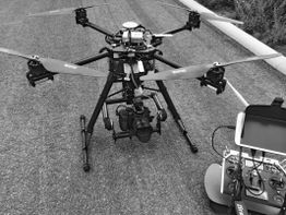 Sechskopter-Drohne mit Kamera auf Asphalt, neben einem Steuergerät im Freien.