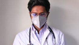 Doctor wearing a face mask and stethoscope, looking down against a plain background.