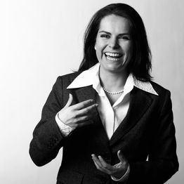 Frau im Anzug lacht und gestikuliert vor neutralem Hintergrund. Schwarz-weißes Foto.