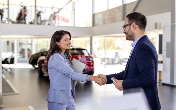 Eine Autohändlerin schüttelt einem Mann die Hand und gratuliert ihm zum Mietvertrag des Autos. Sie stehen in einem Autohaus. Im Hintergrund sind verschiedene Autos und Motorräder zu sehen.