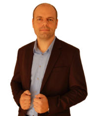 Ein Mann in einem Anzughemd und Sakko schaut in die Kamera, Hände am Revers des Sakkos.