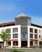 Weißes Bankgebäude mit rotem Dach und Sparkasse-Logo an einer Straßenecke bei klarem Himmel.