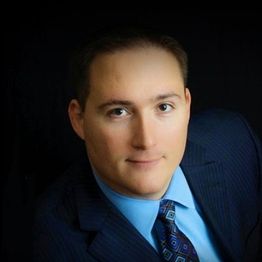 A person in a blue suit and tie with a subtle smile against a dark background.