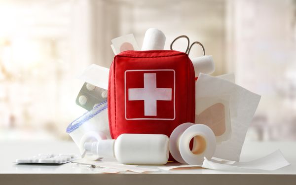 Ein roter Notfallkoffer steht auf einem Tisch. Es schauen Tabletten, Verbände und eine Schere heraus.