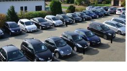 Parkplatz mit vielen Autos in Reihen, umgeben von Grün und Gebäude im Hintergrund.