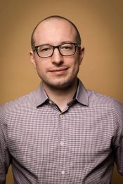 Eine Person mit Brille und kariertem Hemd vor einem braunen Hintergrund.