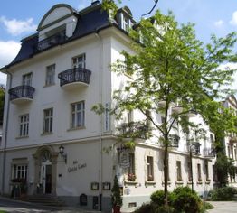 Historisches Gebäude mit Balkonen und Baum im Vordergrund, blauer Himmel im Hintergrund.