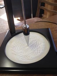 Ein Pendel zeichnet Muster im Sand auf einem quadratischen Tischgerät.