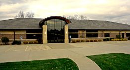 Single-story brick building with large glass entrance and empty parking lot under cloudy sky.