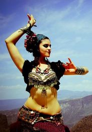 Bailarina con traje colorido y joyas, posa al aire libre bajo un cielo azul claro.