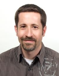 Eine lächelnde Person mit Bart, in einem gestreiften Hemd vor weißem Hintergrund.