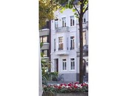 Weißes Gebäude mit Balkon, umgeben von Blumen und Bäumen. Im Hintergrund ein weiteres Gebäude.