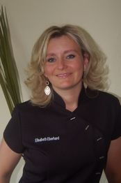 Eine Frau mit blonden Haaren und Ohrringen lächelt in schwarzer Arbeitskleidung.