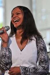 Frau singt lächelnd mit Mikrofon, trägt weißes Oberteil mit gemusterten Ärmeln.