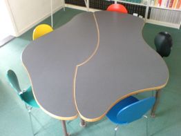 Vier bunte Stühle umgeben einen wellenförmigen, geteilten Tisch in einem Raum mit grünem Boden.