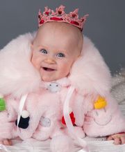 Niedliches Baby in rosa Mantel und Krone, lächelt, umgeben von Kuscheltieren.