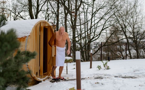 Mann verlässt eine hölzerne Fasssauna in einem verschneiten Garten und trägt ein weißes Handtuch um die Hüfte.