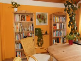 Holztisch, Rattanstuhl, zwei Bücherregale, Pflanzen und orangene Wand in gemütlichem Raum.