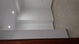 Habitación vacía con paredes blancas y techo con luces empotradas. Suelo gris y puerta de madera.