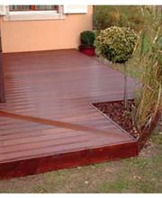 Holzterrasse mit Pflanzenecke vor einem Haus, daneben Rasen und ein paar Büsche.