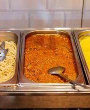 Buffet com três pratos: espaguete, molho vermelho e purê de batatas, com colheres em cada.