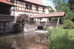 Alte Wassermühle mit Fachwerkgebäude und grünem Umland an einem sonnigen Tag.