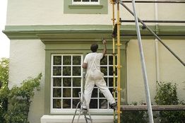 Man painting house window frame green using a ladder and scaffold.