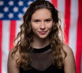 Young woman with long hair smiles slightly, standing in front of a blurred American flag background.