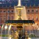 Brunnen im Schlossplatz mit beleuchtetem Gebäude im Hintergrund, zwei Menschen auf einer Bank.
