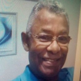 Homem sorridente de óculos, cabelo grisalho, camisa azul, em um ambiente interno com quadro ao fundo.