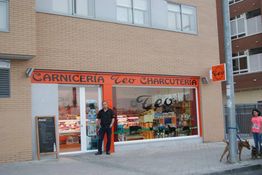 Hombre frente a Carnicería y Charcutería con letrero naranja, junto a una mujer con un perro.