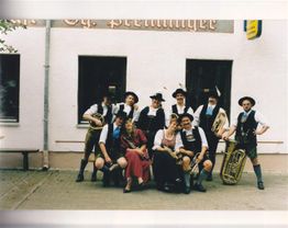 Gruppe von Musikern in traditioneller bayrischer Tracht und zwei Frauen in Dirndln vor einem Gebäude.