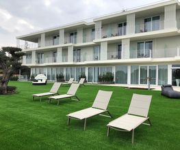 Moderne, mehrstöckige Ferienanlage mit Balkon, Liegestühlen auf grüner Rasenfläche im Vordergrund.