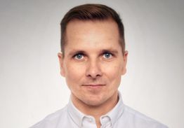 Eine Person in einem weißen Hemd mit neutralem Gesichtsausdruck vor grauem Hintergrund.