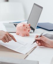 Zwei Personen besprechen ein Dokument am Tisch, mit Laptop und Sparschwein im Hintergrund.