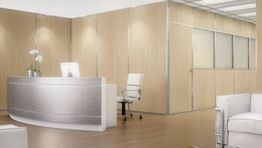 Recepção moderna com paredes de madeira clara, mesa curva, cadeira e orquídea decorativa.