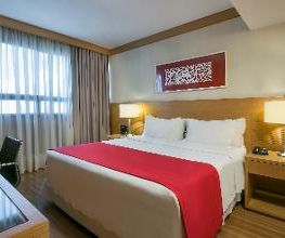 Quarto de hotel com cama de casal, colcha vermelha, luminárias e janela com cortina cinza.
