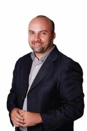 Homem sorridente de terno escuro e camisa clara, com fundo branco.