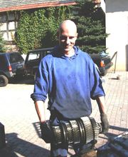 Eine Person hält ein großes Zahnrad, im Freien, mit Autos im Hintergrund.