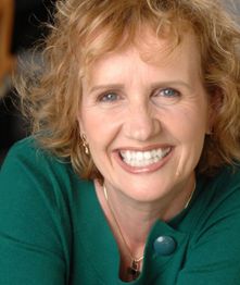 Lächelnde Frau mit lockigem Haar in einem grünen Pullover vor unscharfem Hintergrund.