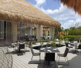 Restaurante al aire libre con techo de paja y mesas negras, rodeado de vegetación tropical.