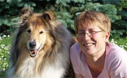 Eine Frau in rosa Shirt lächelt neben einem Collie im Garten.