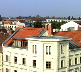 Mehrstöckiges Gebäude mit rotem Dach und Schornstein, umliegende Stadtansicht im Hintergrund.