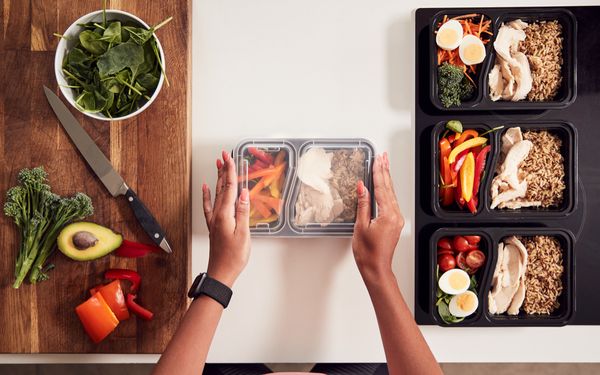 Person bereitet mehrere Meal-Prep-Boxen mit Hähnchen, Reis, Gemüse, Eiern und frischen Zutaten vor.