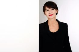 Eine Frau in schwarzem Blazer lächelt und lehnt sich gegen eine weiße Wand.