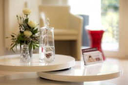 Ein Tisch mit einer Glasvase, Wasserkaraffe und Broschüren, Hintergrund Stuhl und Fenster.