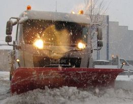 Schneepflug räumt Schnee unter Schneefall von einer Straße, Scheinwerfer leuchten hell.